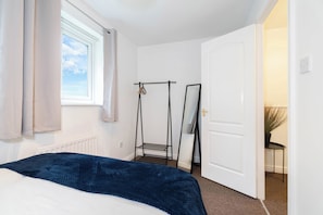 Um quarto com cama, janela com cortinas, radiador, porta e uma pequena mesa com uma planta.