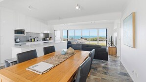 A modern kitchen with white cabinets, a dining area with a wooden table, and a large window offering a view of the ocean. - Seascape at Boomer 2 Ocean Rd, Port Elliot (Port Elliot)
