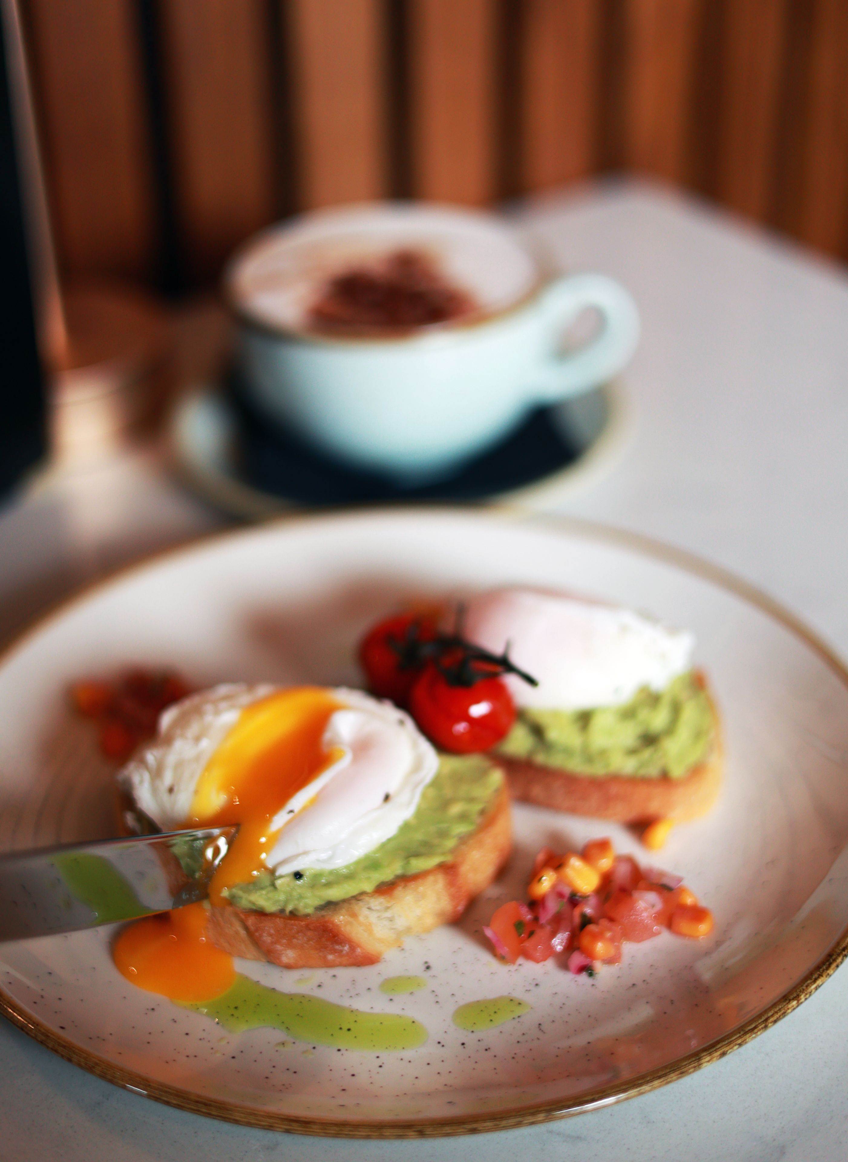 Une assiette avec un œuf poché, de l’avocat et des tomates sur une tranche de pain grillé, et une tasse de café en arrière-plan.