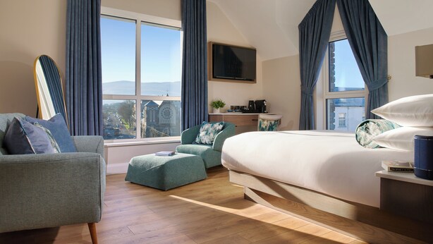 A modern hotel room with a large bed, a seating area with a chair and ottoman, a television, and a view of the cityscape. - The James Hotel (Tralee)