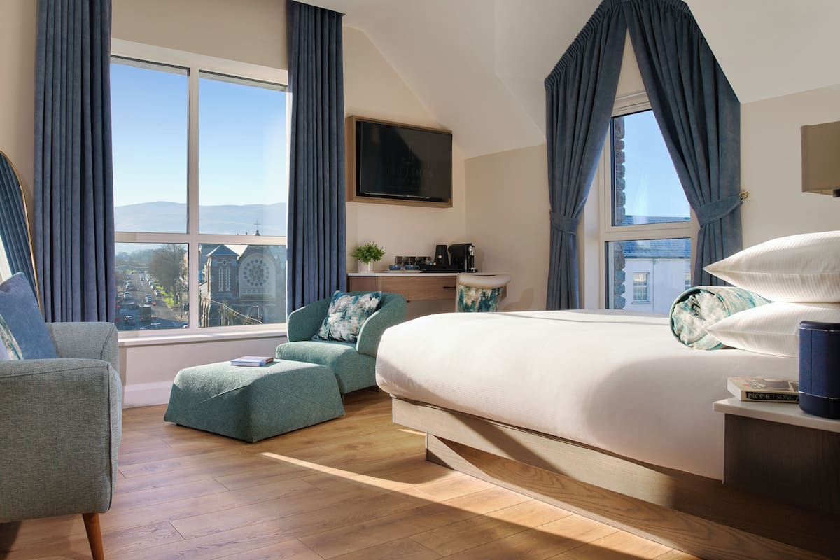 Une chambre d’hôtel moderne dotée d’un grand lit, d’un coin salon avec un fauteuil et un pouf, d’une télévision et offrant une vue sur le paysage urbain.
