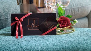 A champagne bottle in a metallic bucket, two wine glasses, and a black gift voucher with a red ribbon on a textured blue sofa. - The James Hotel (Tralee)