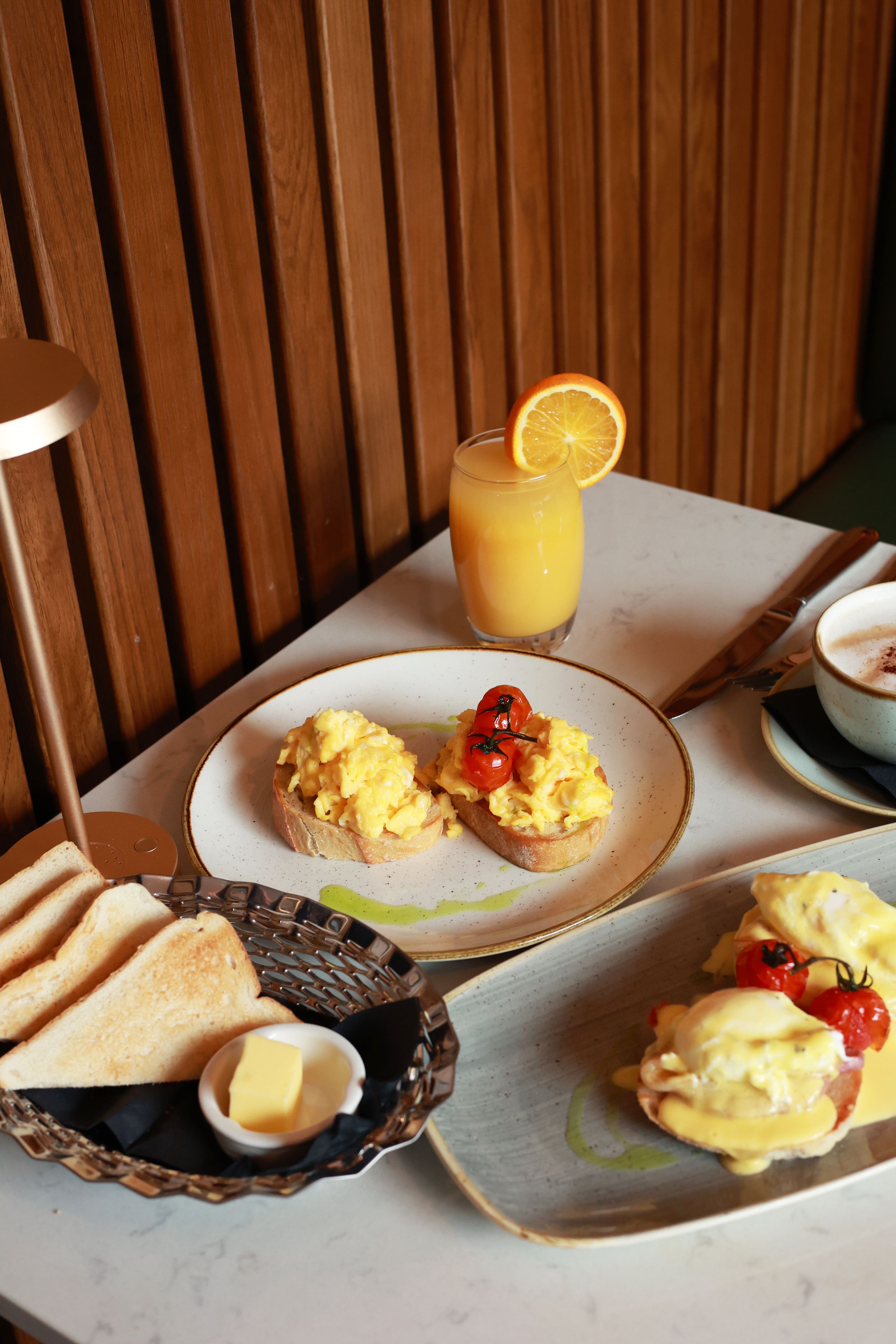 Desayuno con tostadas, huevos revueltos, tomates, un vaso de jugo de naranja y un recipiente de mantequilla.