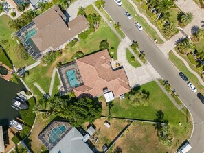 An aerial view of a residential area with a house featuring a swimming pool, a tennis court, and a well-maintained lawn.