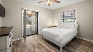 A bedroom with a bed, a ceiling fan, a nightstand, a TV, and a sliding glass door leading to a balcony. - Surfside Beach House | Pvt Pool, Grill, Office (Apollo Beach)