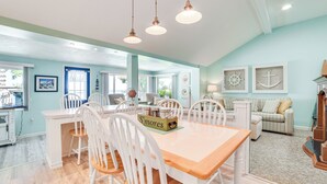 Un amplio comedor con una mesa de madera y sillas blancas, una cocina al fondo y una sala de estar con un sofá a rayas y decoración náutica.