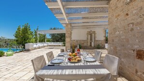 Un espace extérieur aménagé pour les repas, avec une table dressée pour quatre personnes, entourée de chaises en osier. Une piscine est visible au fond, et l’ensemble est couvert d’une pergola.