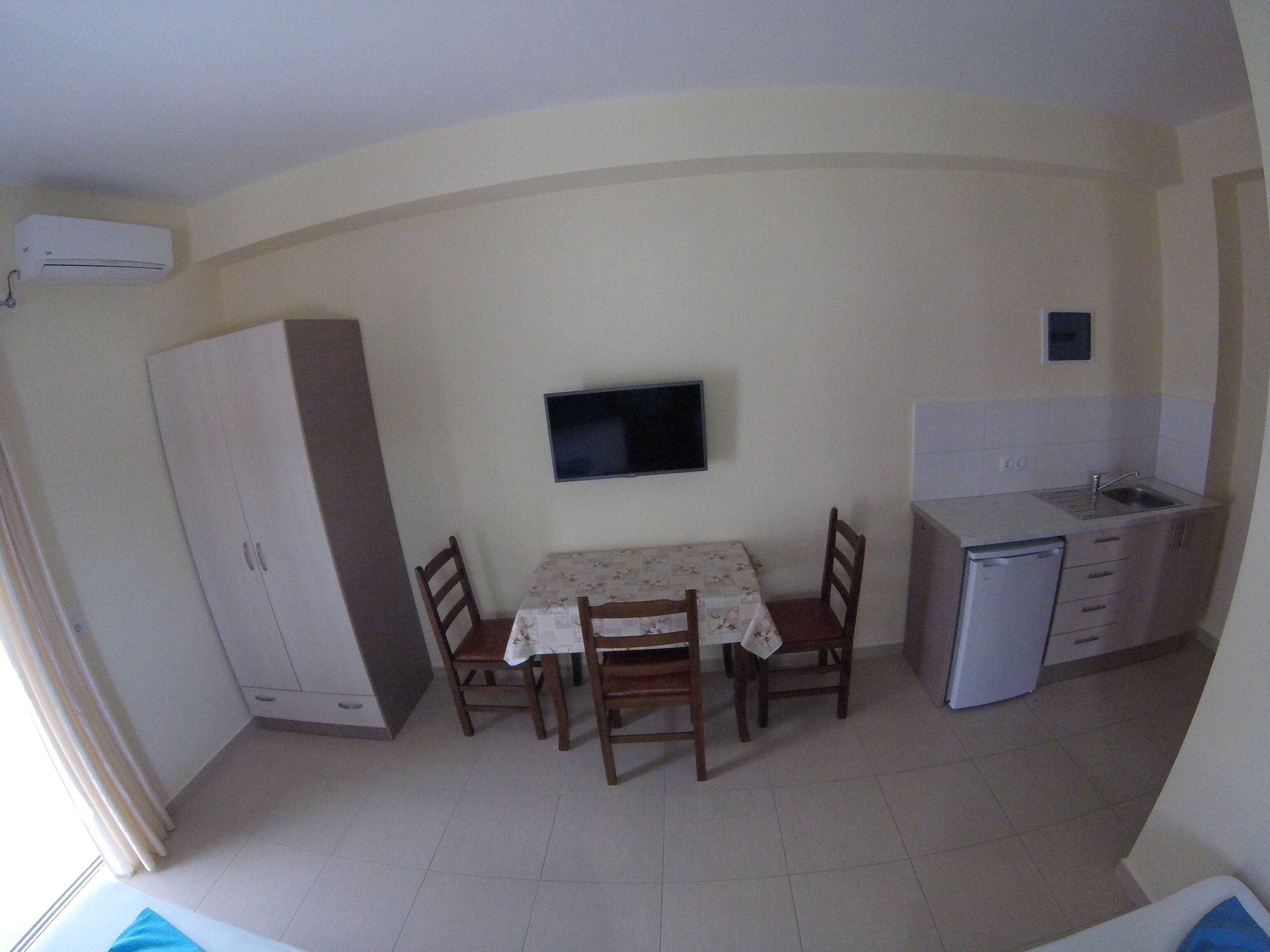 A compact kitchen with a dining table, chairs, a TV mounted on the wall, a small sink, and a refrigerator.