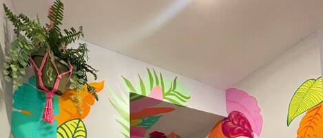 A room with colorful wall murals of tropical leaves and a frog, a potted plant, and a sign with a cheerful message.