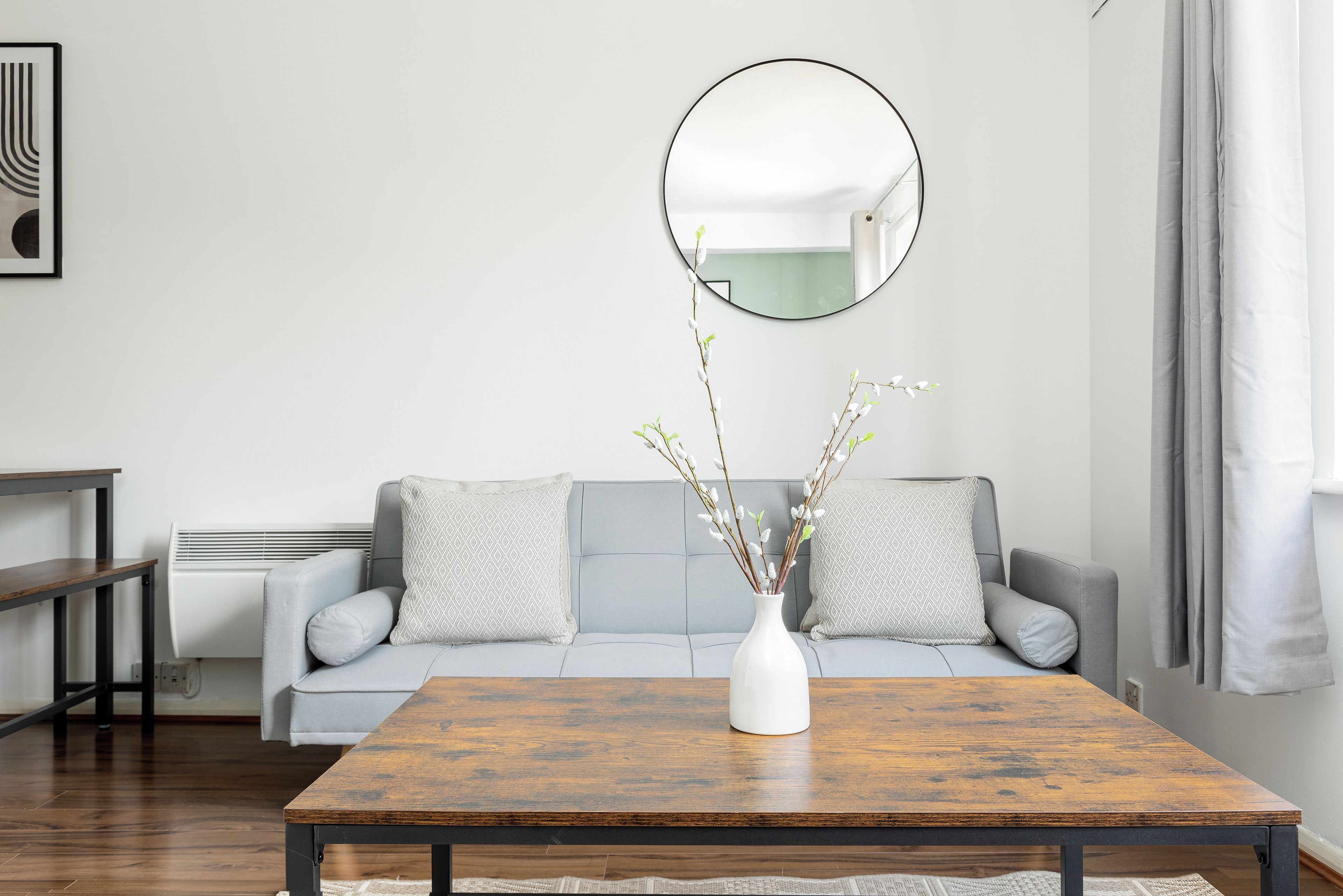A living room with a sofa, a coffee table, a white vase with branches, a round mirror, and a window with curtains.