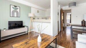 A modern living room with a wooden coffee table, a white vase with branches, a flat-screen TV on a white console, and a minimalist kitchen in the background. - The Bridge Flats (London)