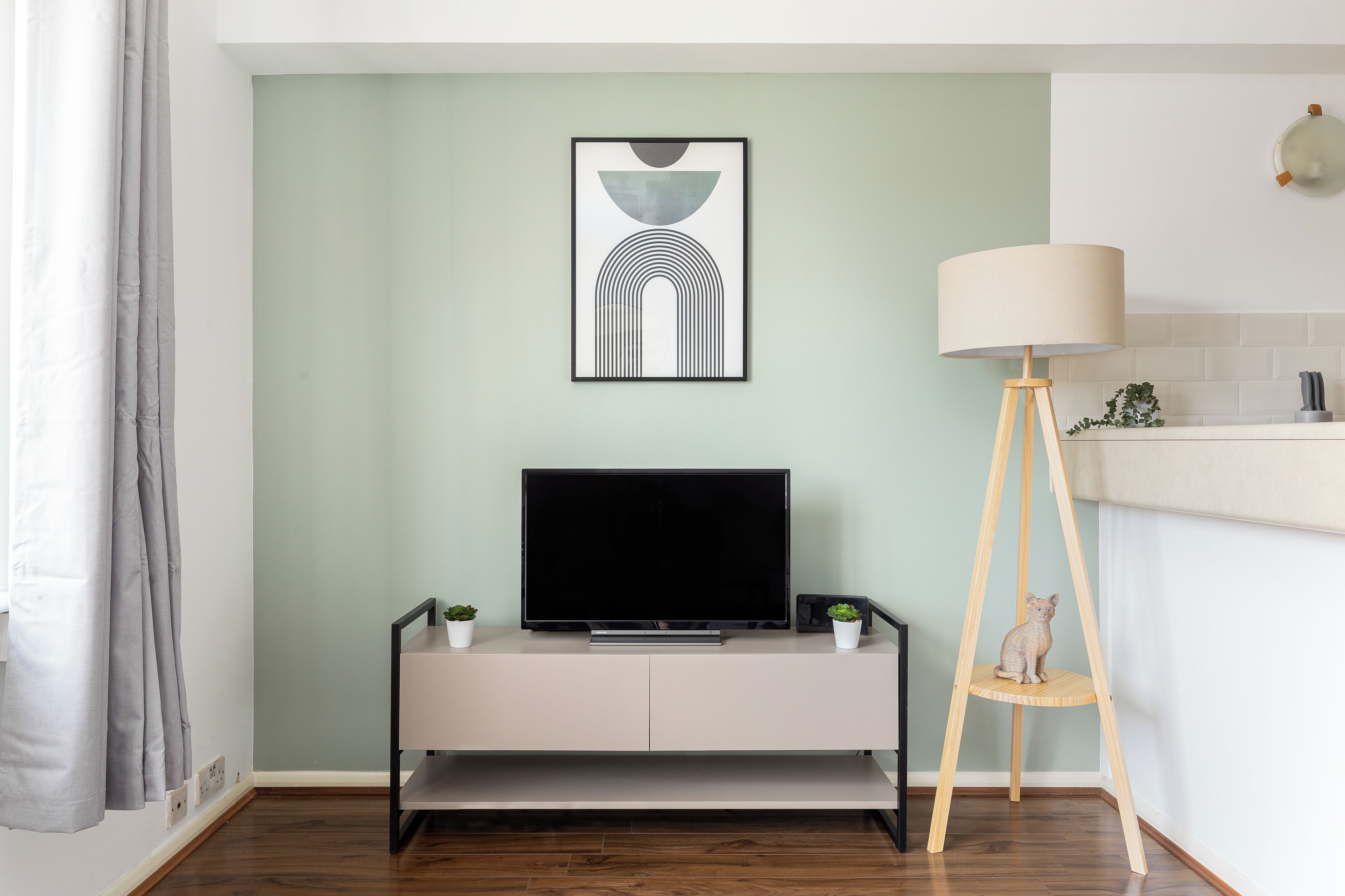 Ein modernes Wohnzimmer mit Flachbildfernseher auf einer schlichten Kommode, einer Stehlampe und einem gerahmten abstrakten Kunstwerk an der Wand.