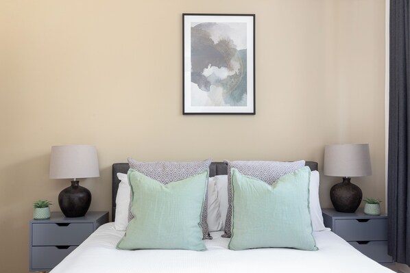 Un dormitorio ordenado con una cama, dos mesitas de noche, dos lámparas y una obra de arte enmarcada en la pared.