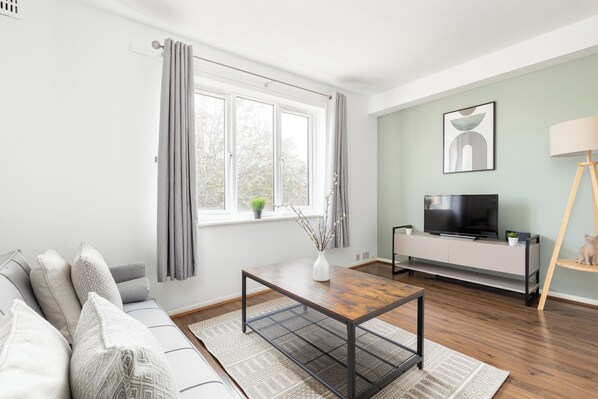 Una sala de estar moderna con un sofá, una mesa de centro, un televisor sobre un mueble blanco y una ventana con cortinas.