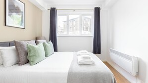 A neatly made bed with pillows, a window with curtains, and a radiator. - The Bridge Flats (London)