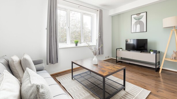 A modern living room with a sofa, a coffee table, a TV on a white stand, and a window with curtains. - The Bridge Flats (London)