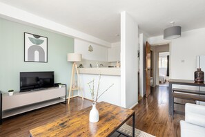 A modern living room with a wooden coffee table, a white vase with branches, a flat-screen TV on a white console, and a minimalist kitchen in the background.
