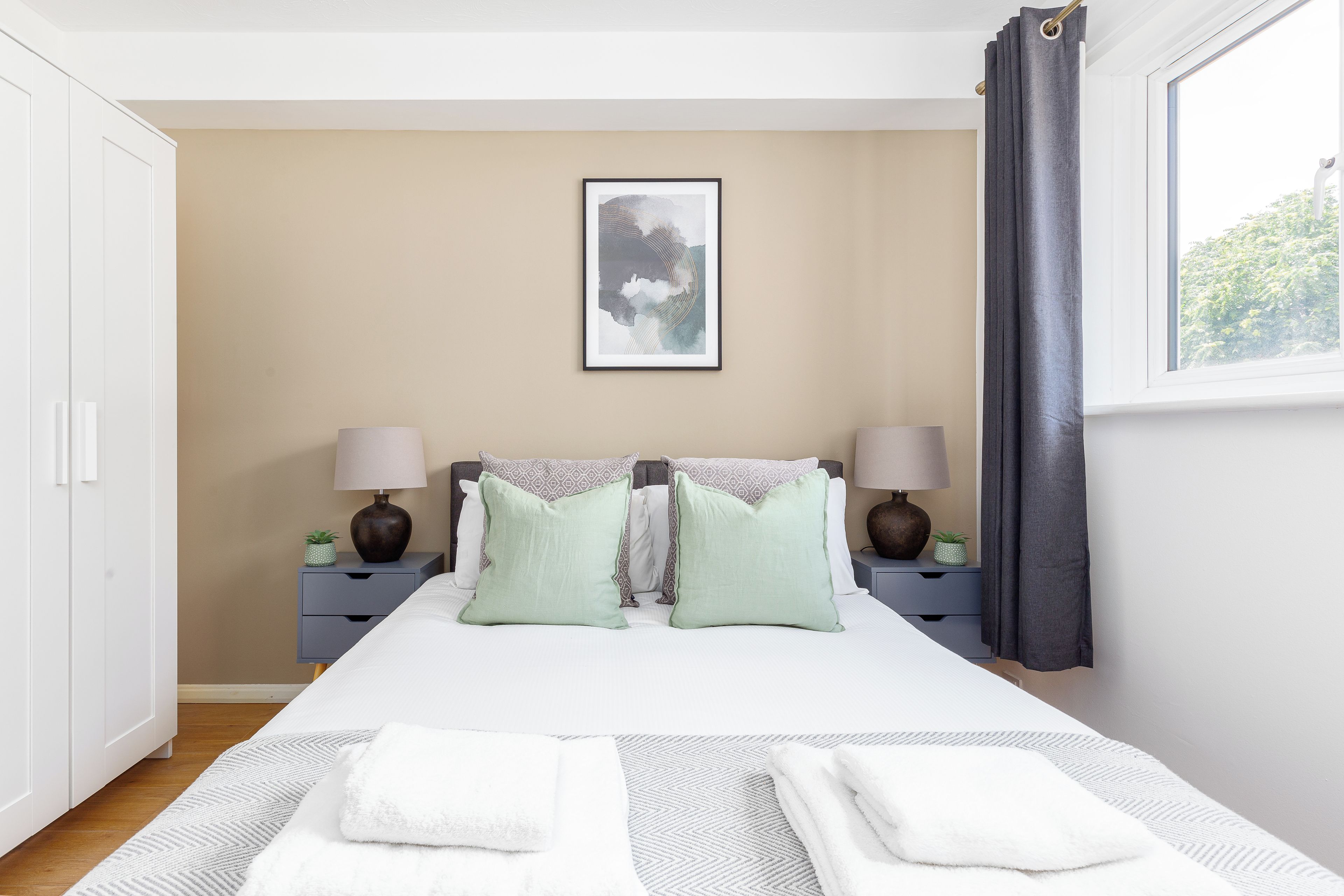 A bedroom with a bed, two bedside tables with lamps, a framed artwork on the wall, and a window with curtains.