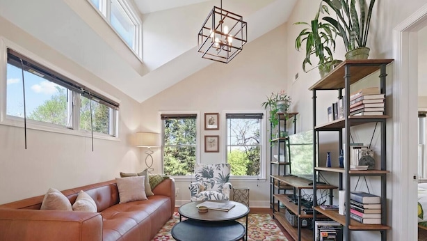 A spacious living room with a brown leather sofa, a round coffee table, and a bookshelf filled with books and decorative items. Large windows allow natural light to fill the room. - Locust Garden, A North Downtown C'ville Guesthouse (Charlottesville)
