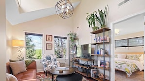 A cozy living room with a sofa, a coffee table, a TV, a bookshelf, and a bed with a floral bedspread. - Locust Garden, A North Downtown C'ville Guesthouse (Charlottesville)