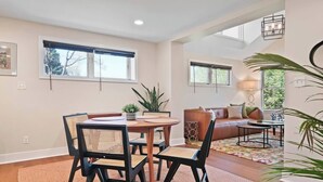 A spacious living room with a wooden dining table and chairs, a sofa, and a coffee table. There are large windows providing natural light and views of the outdoors. - Locust Garden, A North Downtown C'ville Guesthouse (Charlottesville)