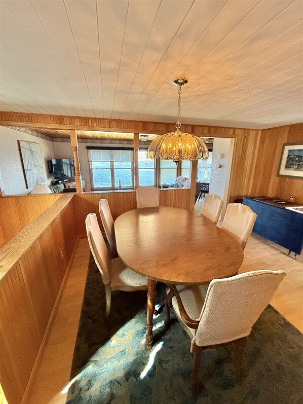 Un espace repas avec une table et des chaises en bois, un lustre et une vue sur la mer grâce à de grandes fenêtres.