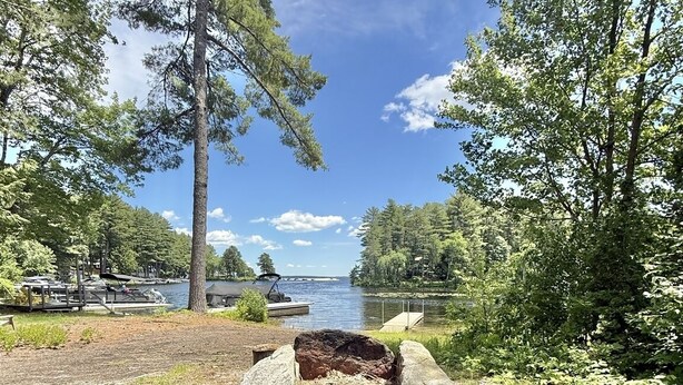 An outdoor seating area with two Adirondack chairs, a fire pit, and a view of a lake with a dock and trees. - WATERFRONT NEWER HOME WITH A DOCK.  4 BEDROOM 2 BATH.   (Sebago)