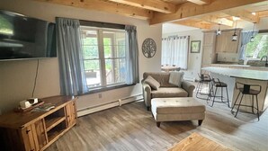 A cozy living room with a wooden ceiling, a flat-screen TV, a sofa, an ottoman, a wooden coffee table, and a kitchen area with a bar counter and stools. - WATERFRONT NEWER HOME WITH A DOCK.  4 BEDROOM 2 BATH.   (Sebago)