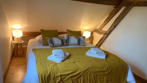 Ein gemütliches Schlafzimmer mit schrägem Dach, einem Bett mit gelber Tagesdecke, zwei Kissen und zwei gefalteten Handtüchern. Auf Holznachttischen stehen jeweils zwei Nachttischlampen.