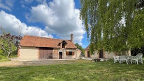 Ein rustikales Bauernhaus mit Ziegeldach, einem Steinsschornstein und einer kleinen Veranda. Vor dem Haus erstreckt sich ein Rasen mit einem Picknicktisch und Stühlen.