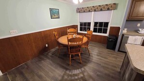 Un coin repas avec une table ronde en bois et des chaises, une fenêtre avec des stores et un espace cuisine doté d’un plan de travail et d’un égouttoir à vaisselle.