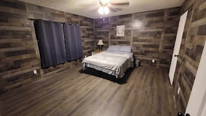Une chambre avec un plancher et des murs en bois, un lit recouvert d’une courtepointe blanche, une table de chevet avec une lampe et un ventilateur de plafond.