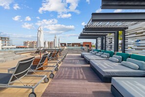 A rooftop lounge area with sun loungers, a pool, and a view of the city skyline.