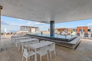 A rooftop bar with a pool, white tables and chairs, and a view of the sea and buildings. - 1BR w/ Rooftop Pool, Bar + City Views | Old Town (Puerto Vallarta)
