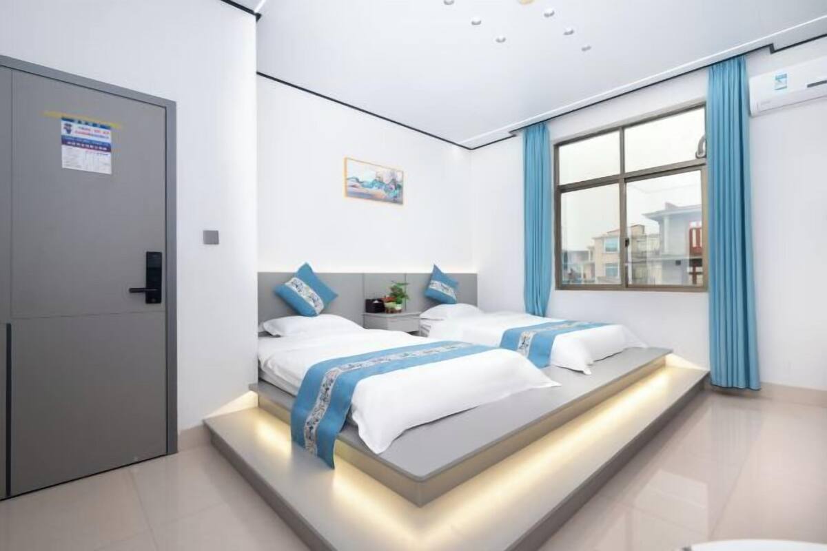 Une chambre d’hôtel moderne avec deux lits, une grande fenêtre avec des rideaux bleus et une vue sur les bâtiments environnants.