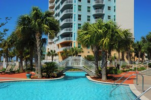 A swimming pool with a bridge, palm trees, and a multi-story building with balconies.