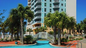 A swimming pool with a bridge, palm trees, and a multi-story building with balconies.