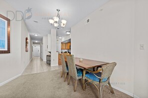 A dining area with a wooden table and wicker chairs, a chandelier, and a kitchen in the background.