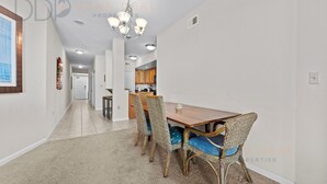 A dining area with a wooden table and wicker chairs, a chandelier, and a kitchen in the background.