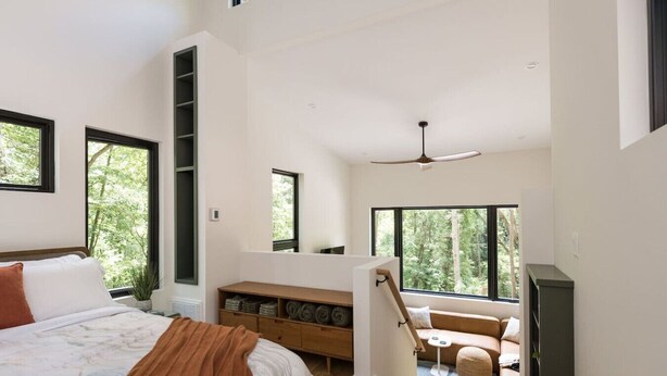 A spacious bedroom with a large bed, a wooden bench, and a view of the outdoors through multiple windows.