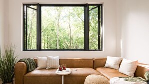 A modern living room with a brown leather sofa, a small white side table, and a large window with a forest view.