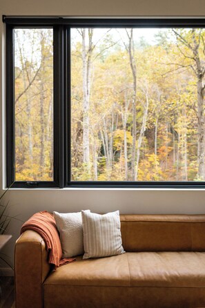 Una habitación con un sofá de cuero marrón, una ventana que ofrece vistas a árboles otoñales y una almohada blanca.
