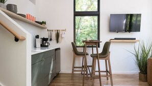 A modern kitchen with a dining area, wooden chairs, a TV mounted on the wall, and a large window.