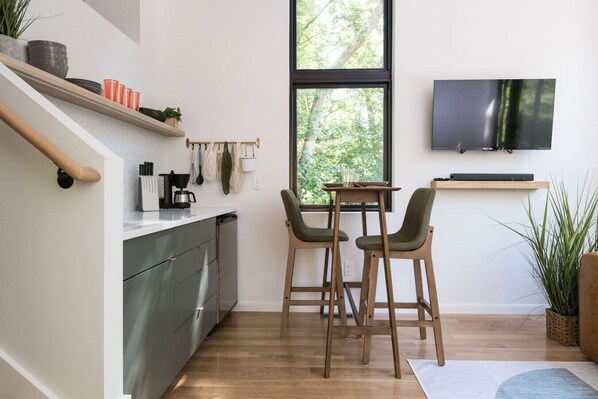 A modern kitchen with a dining area, wooden chairs, a TV mounted on the wall, and a large window. - Modern Tiny Home 4 mile drive to DT w/ Hot Tub (Asheville)