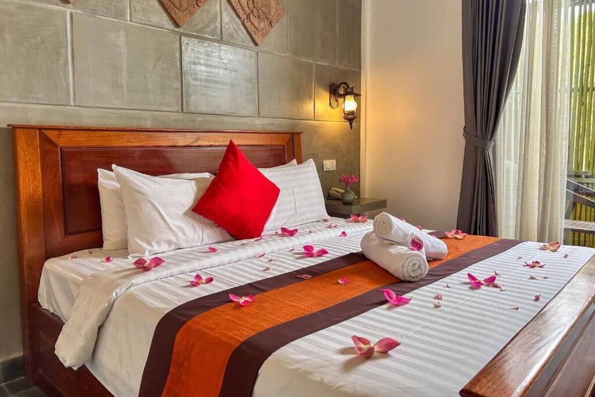 Una cama bien hecha con ropa de cama blanca y marrón, una almohada roja y una toalla enrollada al lado.