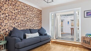 A modern living room with a grey sofa, a wall covered in wooden logs, and a glass door leading to another room. - Glynmar - Sea Glimpses, Hot Tub, Pool Table (Saundersfoot)
