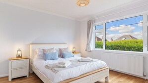 A bedroom with a wooden floor, a white bed with multiple pillows, a nightstand with a lamp, and a window with a view of greenery and a house. - Glynmar - Sea Glimpses, Hot Tub, Pool Table (Saundersfoot)