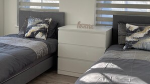 Une chambre avec deux lits, chacun doté d’une tête de lit grise et d’oreillers assortis. On y trouve une table de chevet blanche sur laquelle est posée une petite pancarte portant l’inscription « chez soi ».
