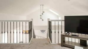 Une pièce spacieuse avec une grande télévision posée sur un meuble en bois, un escalier avec des balustrades noires et un luminaire au plafond.