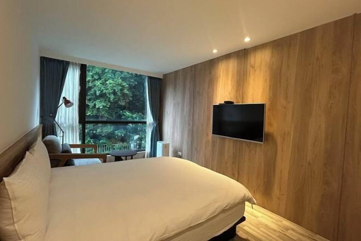 A hotel room with a bed, a TV mounted on the wall, a window with curtains, and a view of greenery outside.